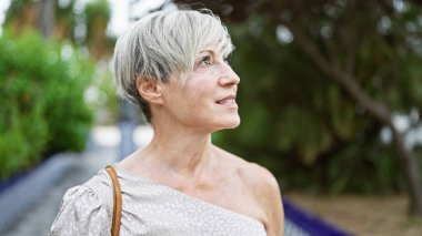 Beyaz saçlı, güzel bir gülüşü olan olgun İspanyol kadın yeşil bir parkta doğanın tadını çıkarıyor..