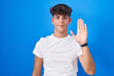 Mavi arka planda duran İspanyol genç merhaba diyor mutlu ve gülümsüyor, dostça karşılama jesti 