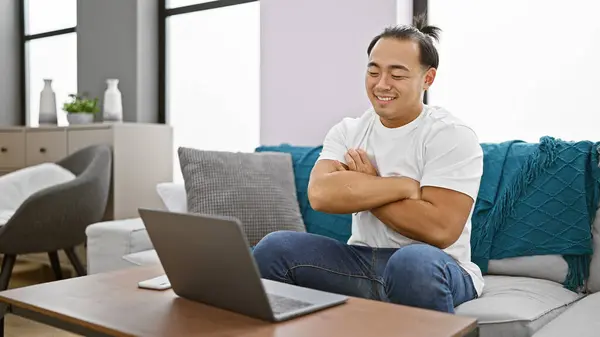 Cheerful young chinese man relaxed indoors, sitting in his living room\'s interior with arms crossed, happily using technology on his laptop at home.