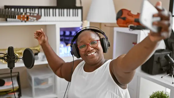 自信的非洲裔美国女音乐家 戴着辫子和眼镜 在一家音乐工作室的智能手机上自拍时微笑着 — 图库照片