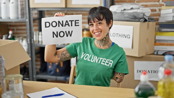 Hispanic woman with amputee arm volunteer smiling confident holding donate now message at charity center
