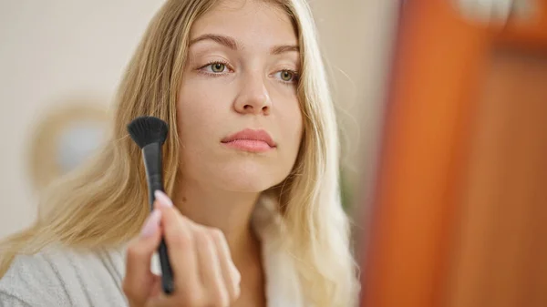 Young blonde woman looking on mirror applying makeup looking on mirror at bedroom