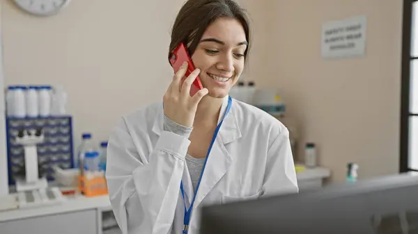 A smiling young woman in a lab coat talking on a phone in a laboratory setting.