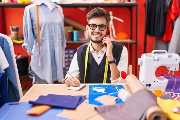 Young hispanic man tailor talking on smartphone writing on notebook at atelier