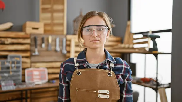 年轻迷人的金发碧眼的女木匠戴着安全眼镜 在她忙碌的木匠作坊里表现出严肃的专业精神 — 图库照片