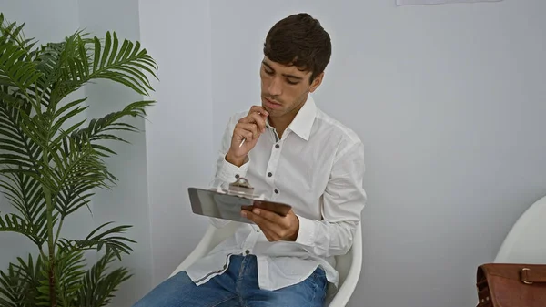 Attractive young hispanic man, engrossed in serious thought, meticulously taking notes while sitting in the waiting room, prepping for a business meeting or job interview.