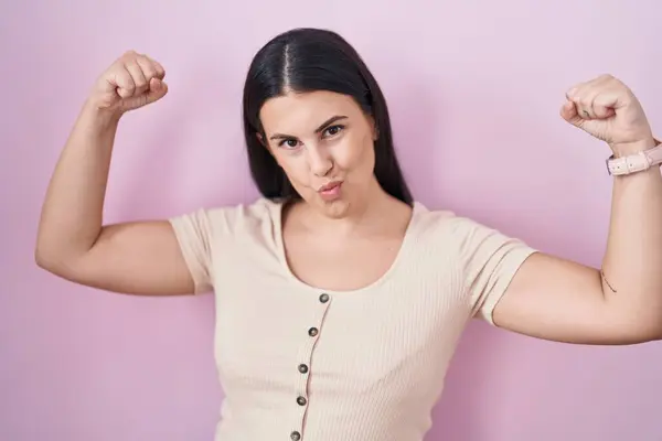 Young hispanic woman standing over pink background showing arms muscles smiling proud. fitness concept.