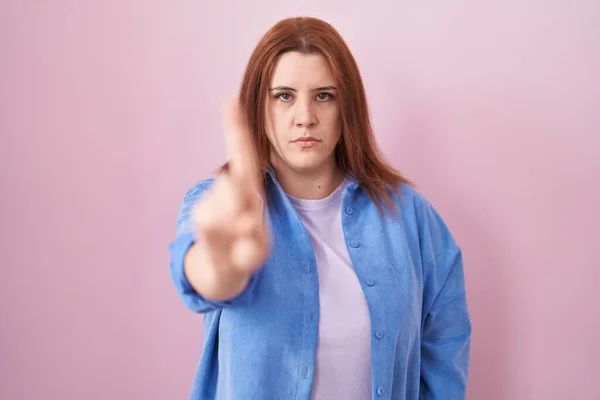 指と怒りの表情を指してピンクの背景の上に立っている赤い髪の若いヒスパニックの女性 ジェスチャーを示さない — ストック写真