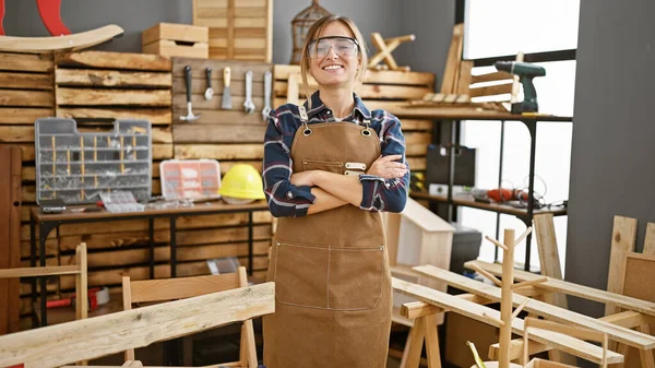 年轻而自信的金发碧眼的木匠女人 戴着眼镜 交叉着双臂 骄傲地站在木工车间里 — 图库照片