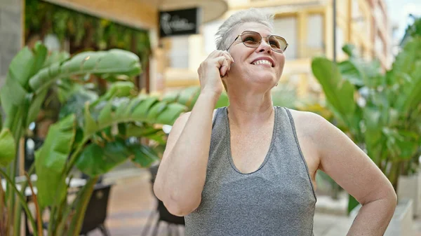 Orta yaşlı, kır saçlı, gülümseyen, kendine güvenen bir kadın parkta akıllı telefondan konuşuyor.