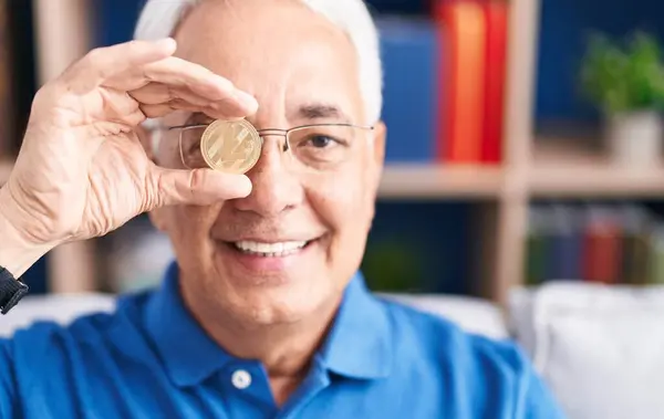 Middle age grey-haired man holding litecoin crypto currency over eye at home