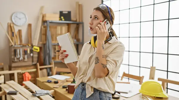 A young woman multitasking in a carpentry workshop, talking on the phone while holding a notebook.