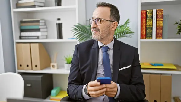 Confident middle age man with grey hair, happily working at his office job. this successful business worker uses his smartphone while sitting at his desk, exuding positivity.