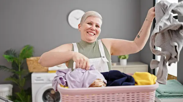 Middle age grey-haired woman smiling confident throwing clothes on air at laundry room