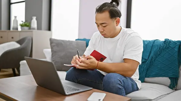 Handsome young chinese man, relaxing indoors on a sofa at his home. focused, he\'s using his laptop, taking notes with serious concentration, surrounded by the comfort of his living room.