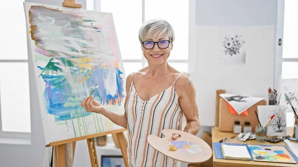Mature woman artist smiling in a studio with a colorful painting, palette, and glasses.