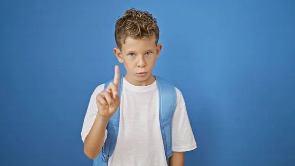 可爱的金发男生 一个聪明的小孩站在那里 严肃地表示否定 用他的手指说不 孤立的蓝色墙壁衬托突出了他的书包 — 图库照片