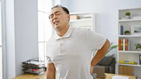 Worried young chinese man worker suffering serious backache, touching aching back, standing unhappily in office indoors. pain from work injury keeps handsome employee from professional tasks.