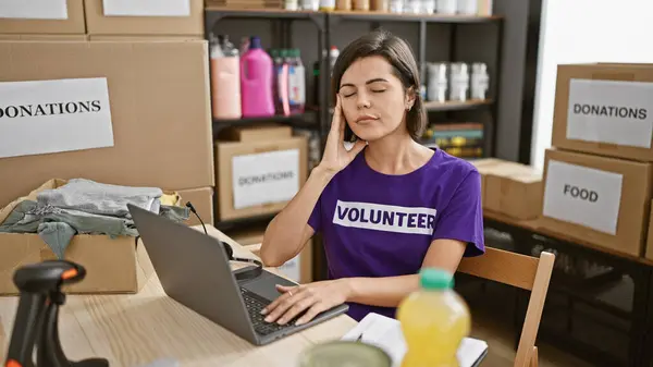 Exhausted young hispanic woman volunteer, with beautiful hairstyle and short hair, feeling overwhelmed while working on her laptop at a bustling charity center, supporting her community