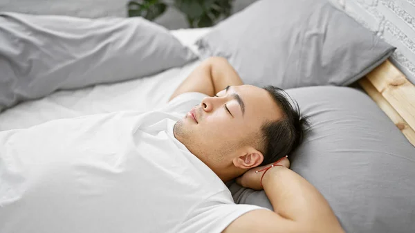 Handsome young chinese man relaxedly lying on a comfy bed, hands behind head, deep in thought in a cozy bedroom, enjoying the morning relaxation at home.