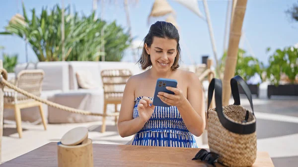 Young hispanic woman smiling happy using smartphone sitting on the table at sunny restaurant terrace