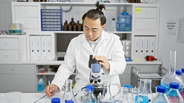 Handsome young chinese scientist man delving into biology with microscope, writing analysis notes at an indoor lab, fully immersed in medical research.