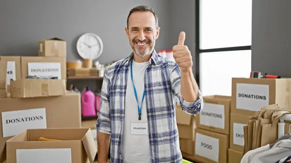 Confident middle-aged man with grey hair enjoys volunteering, smiling and thumbing up at the busy charity center