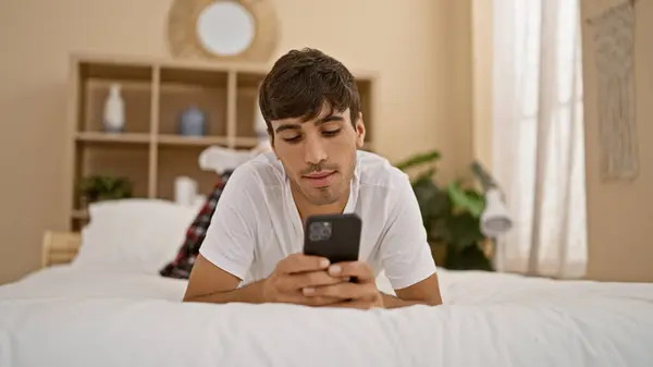 Handsome young hispanic man in pyjamas lying comfortably on bedroom\'s bed, intently using smartphone for morning texting, portraying a relaxed lifestyle, indoors.
