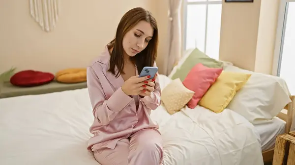 居心地の良い寝室でベッドに座っている間 スマートフォンを使用してパジャマの若いブルネットの女性 — ストック写真