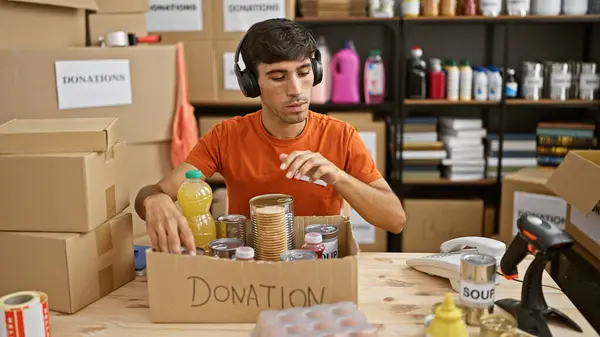 Warm-hearted young hispanic man volunteering at a charity center, confidently packing food into cardboard boxes whilst listening to uplifting music