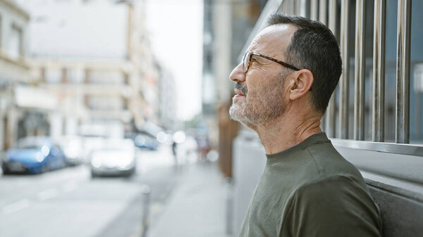 Serious, middle age man with grey hair, glasses, and a beard stands outdoors. as he looks up, his concentrated expression hints his thoughts are lost in the urban city street.