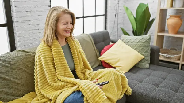 黄色い毛布で覆われた屋内で微笑む白人女性 リモートを保持し 明るい部屋で灰色のソファーに座っている — ストック写真