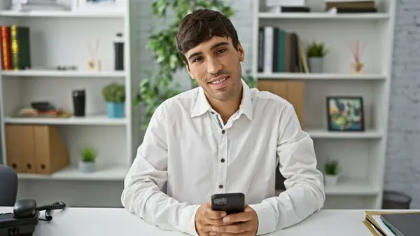 Working with joy, a confident young hispanic man enjoys his work at the office, handsomely typing messages on his smartphone while sitting at his desk, exuding success and positivity.