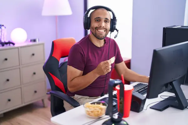 Young latin man streamer playing video game eating chips potatoes at gaming room