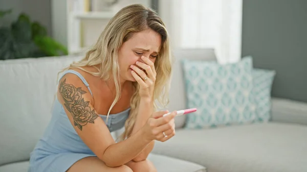 Young blonde woman holding pregnancy test crying at home