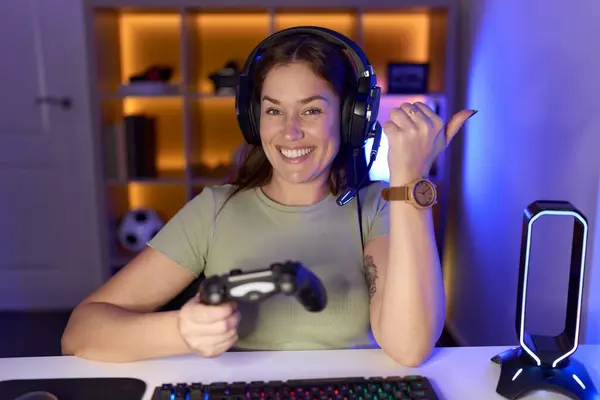 ハンドと親指で背中を指すヘッドフォンを着てビデオゲームをしている美しいブルネットの女性は自信を持って微笑みます — ストック写真