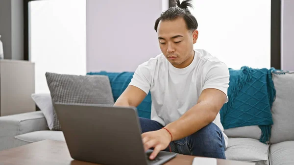 Handsome young chinese man, focused and concentrated, working online using laptop while sitting on sofa indoors. delving deep into the world of internet technology at his apartment\'s living room.