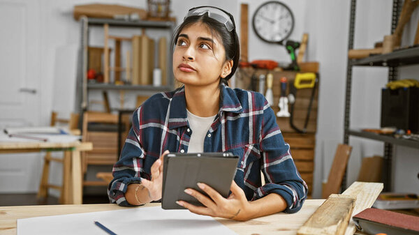 Созерцательная индианка с длинными волосами держит планшет в деревообрабатывающей мастерской, окруженный инструментами и древесиной.