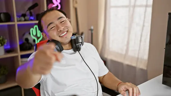 Handsome young chinese guy, a lively digital gamer and streamer, pointing at the camera in a dark gaming room, amidst the thrill of nightly gaming, sitting with a console, headset on