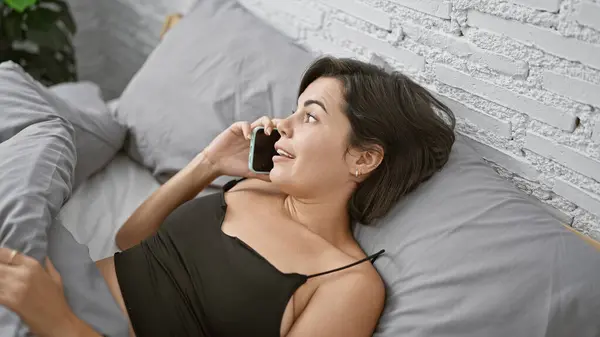 Confident young hispanic woman with a gorgeous hairstyle, happily chatting away on her smartphone, lying relaxed on her comfy bed amidst fluffy pillows and soft blankets in her cosy bedroom