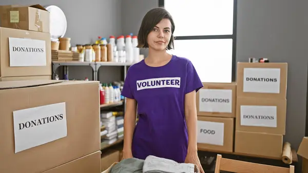 Captivating portrait, young, beautiful hispanic woman, a dedicated volunteer, standing with a serious face amidst the heart of charity work at a busy community center