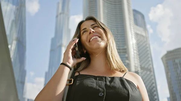 A smiling young woman talks on her phone in front of the towering skyscrapers of dubai under a blue sky.