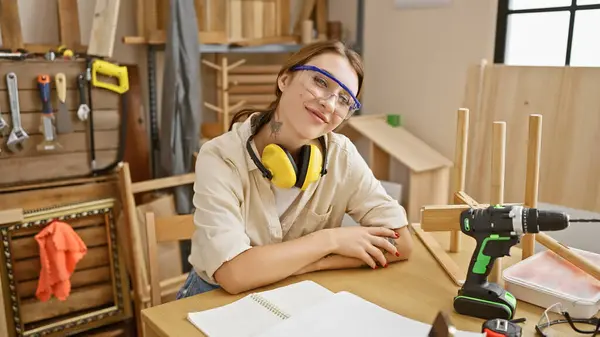 Młoda Kobieta Okularach Słuchawkach Uśmiecha Się Warsztacie Stolarskim Otoczona Drewnem — Zdjęcie stockowe