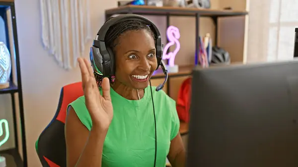 Joyful african woman in a gaming room wearing headphones and waving indoors