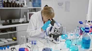 Ciddi, uyumlu sarışın bir bilim adamı, orta yaşlı bir kadın, laboratuarda çalışıyor, mikroskop kullanarak tıbbi araştırmalara katılıyor, notlar alıyor, hayat değiştiren bir keşifle ilgili bilgi veriyor..