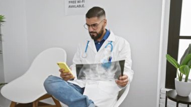 Gözlüklü sakallı adam klinikte akıllı telefonla birden fazla iş yaparken röntgen analizi yapıyor..