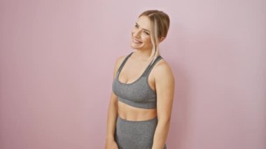Sportif ve kendinden emin, spor kıyafetli genç sarışın bir kadın rahat bir poz veriyor, doğal bir gülümsemeyle yan tarafa bakıyor. Pembe izole arka plan için fazla havalı..