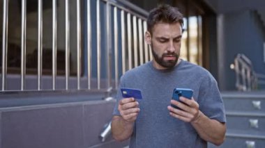Kendine güvenen, gülümseyen İspanyol genç adam, şehir caddesinde duruyor, akıllı telefon ve kredi kartı kullanarak online alışverişin tadını çıkarıyor, kentsel yaşam tarzını kucaklıyor.