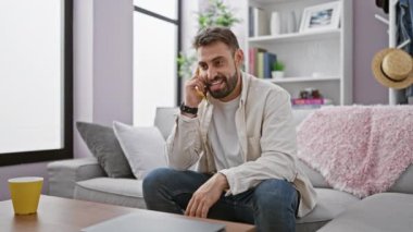 Neşeli, genç İspanyol bir adam kanepeye oturmuş, rahat bir evde, neşeli bir şekilde telefonla sohbet ediyor, pozitiflik saçıyor.