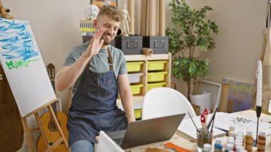 Mavi gözlü, sakallı beyaz bir adam yaratıcı bir stüdyoda dizüstü bilgisayarla çalışırken gülümsüyor..